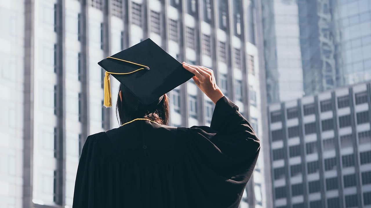 Student in cap and gown in front of city buildings